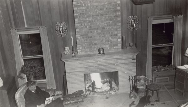 Elevated view of Ernst Conrad Schmidt (1893-1977), grandson of Chicago brewer Conrad Seipp, sitting in an easy chair reading in his high ceilinged, wood paneled living room. A fire blazes in the fireplace. His home was referred to as the Baker house or "the farmhouse" on the Black Point Estate.