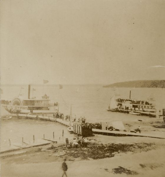 Elevated view from shoreline of two side wheeled steamboats, the "Lucius Newberry," left, and the "Lady of the Lake," docked at Geneva Lake. Other smaller boats are also tied to the piers and sailboats are seen on Geneva Lake in the background. Cord wood is stacked on the shore near the closer pier. People area standing on both piers.