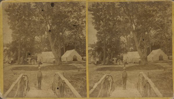 Stereograph view from footbridge with rustic railings of a small boy wearing a large hat standing at the end of the footbridge. A group of adults and another boy stand or sit in the background near several tents under trees.