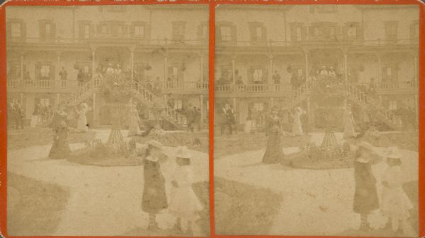 Stereograph of the large hotel at Kayes Park on the south shore of Geneva Lake. There are two girls standing in the foreground, with well-dressed women in the formal garden behind them. Men and women are posing on the double central staircase and two-story porch at the front of the hotel.