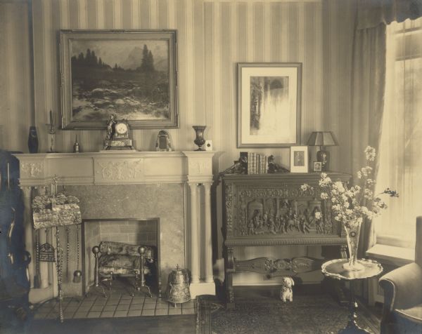 The fireplace in the living room of the Edward Petersen house features a classical revival surround and mantel. A small stuffed toy dog sits under a carved cabinet next to the fireplace. There is a large landscape painting above the fireplace.