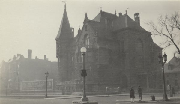 View across street towards a boy and girl standing on the street corner across from the granite mansion built for Conrad Seipp at 3300 South Michigan Avenue. The home features several chimneys and a tower with turret. There are ornamental ironwork spires on the roof. A billdboard advertising "De Luxe Line" stationary products stands incongruously in a vacant lot next door on the left. The house was designed by Adolph Cudell and was completed in 1888. Cudell also designed Seipp's summer home at Black Point on Geneva Lake, which was built at the same time. Seipp died in 1890 but his widow, Catherine Orb Seipp continued to live in the house until her death in 1920. The mansion was torn down in 1933.