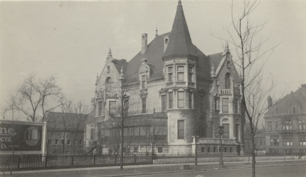 View across streets towards the tower with a steeply pitched roof which anchors the southeast corner of the Conrad Seipp mansion at 3300 South Michigan Avenue. There is a south facing conservatory on the first floor and a porch facing Michigan Avenue. Stone spires adorn the roof of the granite house. A portion of a billboard is on the left.
