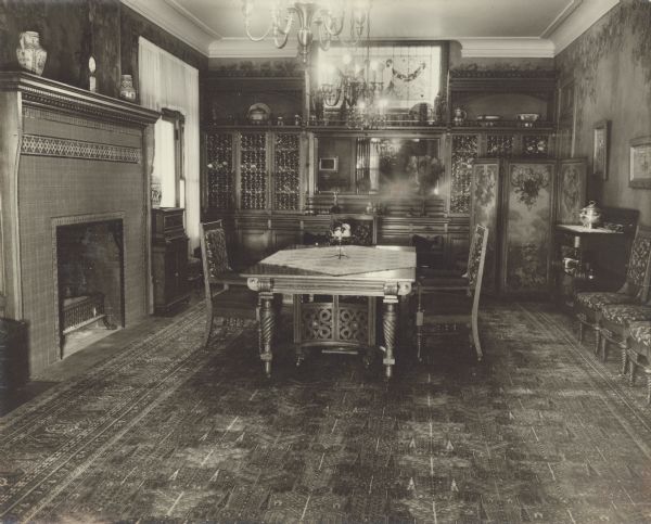 An ornate dining table with matching chairs anchors the dining room at Black Point. There is a stained glass window at the far end of the room over a built-in china cabinet with mirror. A three panel folding screen is on the right. On the floor is an oriental carpet. Stenciled or painted leaves and vines decorate the walls. There is a fireplace with tall tile surround on the left.