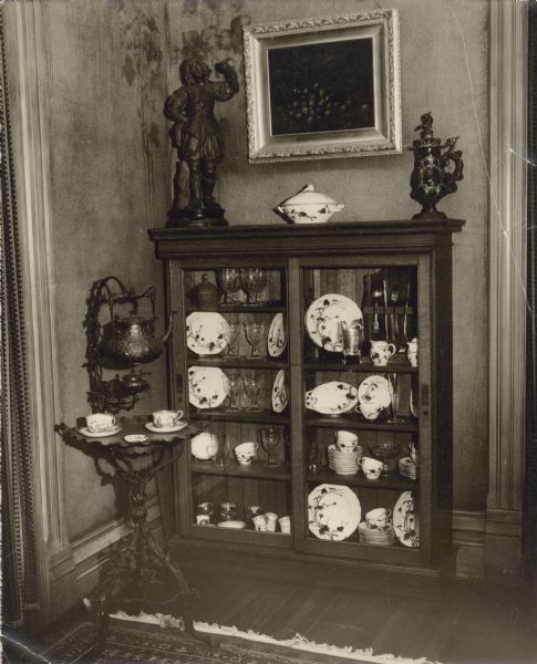 A view of the southeast corner of the dining room in the main house at Black Point, showing a china cabinet and tea table. The cabinet holds pieces of Catherine Seipp's china. A carved wood sculpture of a cavalier stands on top of the cabinet and a still life painting hangs on the wall. The ornate tea table with attached hanging kettle and warming lamp is on the left. According to information on the reverse of the photograph, the table was "from World's Fair 1893." There are decorative leaves and vines painted or stenciled on the walls.
