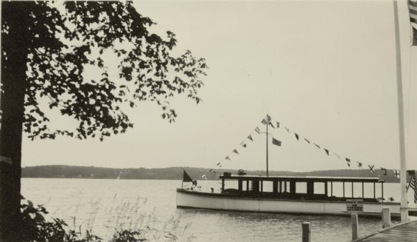 View from shoreline of the motor launch "Nepenthe," owned by Dr. Otto L. Schmidt, resting at the pier at Black Point on Geneva Lake. The boat is "dressed" with International Code flags displayed from its mast, appropriate for July 4.