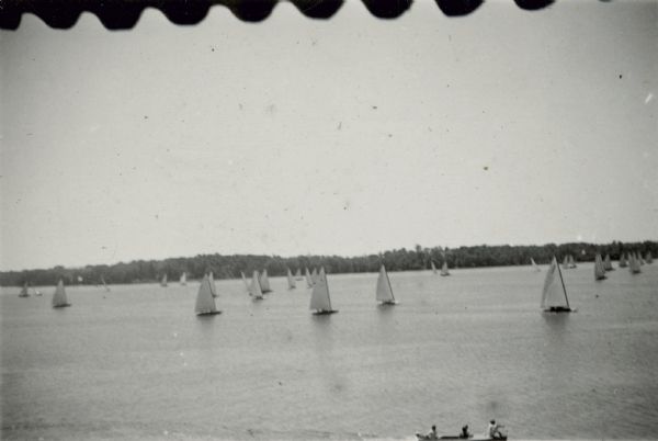 Elevated view across water of sailboats on Geneva Lake. There are people on a boat with an outboard motor in the foreground. The scalloped edge of an awning is along the top in the foreground.