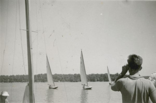 A young man in the foreground, possibly Ernst Schmidt, stands looking out at sailboats on Geneva Lake. In the foreground on the left a woman is on a sailboat, and another person stands next to Ernst on the right. Schmidt was the grandson of Conrad Seipp, who was a Chicago brewer and an owner of Black Point Estate.