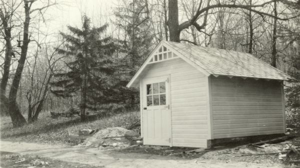 A small, simple wood frame building stands along the brick walkway near the shore of Geneva Lake at Black Point. There is lattice trim under the eaves on the gable end and exposed rafter tails. Construction debris litters the ground. On the reverse of the photograph is written "gas house."