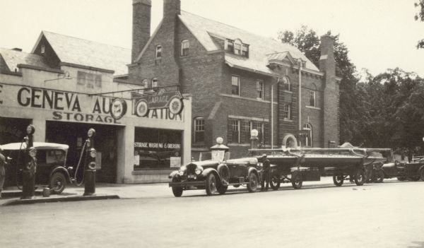 View across street towards a convertible automobile with a fender-mounted spare tire parked in front of the Lake Geneva Auto Station on the south side of the 800 block of Main Street. The car is towing a racing yacht on a trailer. There is a man standing at the rear of the car. Another car is parked near the station's gas pumps. A "Garage" sign in the shape of an automobile hangs over the sidewalk. The 1908 YMCA building at the corner of Main Street and Wrigley Drive is seen in the background; a YMCA sign hangs above the side entrance. The brick building has several large chimneys.