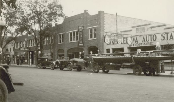View across street towards a convertible automobile with a fender-mounted spare tire parked in front of the Lake Geneva Auto Station on the south side of Main Street. It is pulling a racing yacht on a trailer. A "Garage" sign in the shape of an automobile hangs over the sidewalk; next to it a smaller sign reads "Authorized Buick Service." A Buick sign is also mounted on the front of the adjacent two-story building. The building further to the left, identified by a sign as the Main St. Garage, also has a sign for a jeweler on it. There are gas pumps in front of the Main St. Garage and the Lake Geneva Auto Station.