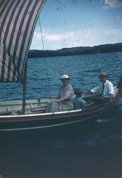 Alma Schmidt Petersen controls the sheets on a small sailboat. Her husband, William F. Petersen, sits aft. The child between them is unidentified. The sail has red and white stripes. In the far background is a tree-lined shoreline. The boat, the "Rhana," was a fishing boat brought from Norway to Lake Geneva in 1937.