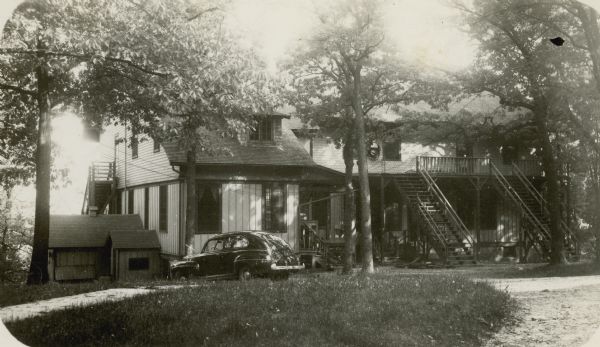 View across lawn towards a car parked in front of the main building at Holiday Home Camp. The rambling wood frame building has several sets of stairs leading to the second story. Geneva Lake is in the background.