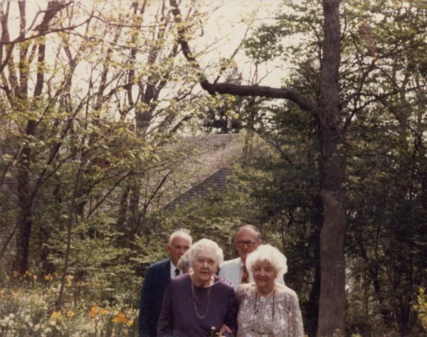 Alma Schmidt Petersen, left, walks arm and arm with her sister, Clara Theresa (Tessa) Reese on the grounds of Black Point Estate.  Behind them are two of Alma's sons, Conrad, left, and Edward Petersen.  Spring flowers are in bloom and the trees are leafing out. In the background is the house of Alma's third son, William O. Petersen.