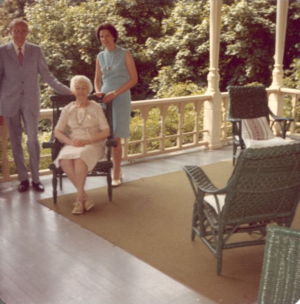 Alma Schmidt Petersen, owner of Black Point Estate, poses seated in a green wicker chair on the porch of the main house. Standing behind her on the left and right is an unidentified man and woman.