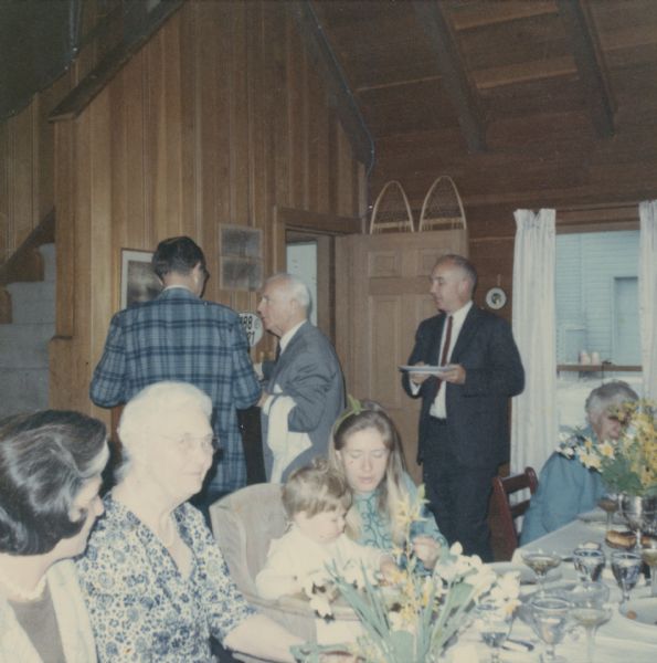 Members of the Petersen family gathered for a meal at the farmhouse, also referred to as the Baker house, at Black Point Estate. The room is open to the rafters and the walls are wood paneled. A typed label on the reverse of the photograph reads, "Seated Judy Bartholomay, Alma Petersen, Baby Edward Goltra, Catherine Goltra, Katherine Lefens.  Standing Edward Petersen, Hans Reese, Wm. O. Petersen at the farm house April 27, 1968." Ernst Schmidt, Alma Petersen's brother, used the farmhouse as a second home.