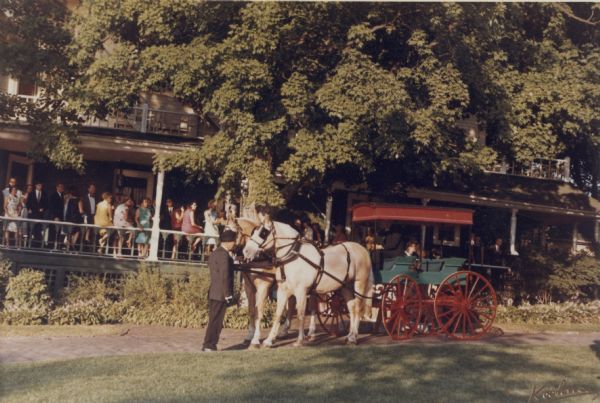View from lawn of a man holding the harness of a team of two horses hitched to an open carriage on the brick drive at Black Point. A large group looks on from the porch of the main house.