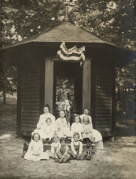 Ernst Schmidt poses in the doorway of the playhouse on the grounds at Black Point Estate. He is wearing an exotic native American style costume adorned with feathers and small furs. Two women sit on either side of the upper step. The woman on the left is unidentified; on the right is Bertha Luehr, nanny to the Schmidt children. The children are, top step from left, Elsa Bartholomay, Alma Schmidt, Katherine (Kate) Bartholomay, unidentified. The children seated on the ground are, from left, Clara Theresa (Tessa) Schmidt, Henry Bartholomay, Fred Stauffer, and Allen Stauffer. There is a horse pull toy on each side of the children. The Bartholomay and Schmidt children were the grandchildren of Conrad and Catherine Seipp, who established Black Point Estate.