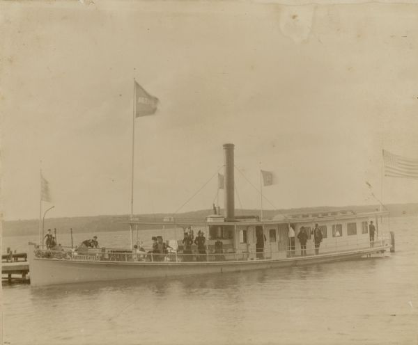 View across water towards men and women in fine clothes posing on the deck of the steamer <i>Arthur Kaye</i>, which is docked at a pier on Geneva Lake. Named for its owner, the boat carried the Kaye family members and clients to Kayes Park Resort on the south shore of Lake Geneva. The ship burned and sank in 1898.
