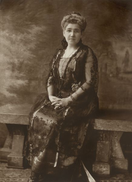 Full-length seated portrait of Wilhelmina Petersen, mother of William F. Petersen M.D. She is wearing the dress that she wore to the wedding of her son to Alma Schmidt, granddaughter of Chicago brewer Conrad Seipp. She is sitting on a stone bench in front of a painted backdrop.