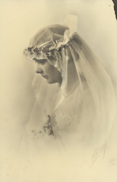 Head and shoulders studio portrait, in profile, of Alma Catherine Schmidt (1894-1989) on the day of her wedding to William F. Petersen M.D. Her veil is secured by a wreath of flowers; only a small portion of her bouquet is visible. She wears a single strand of pearls.