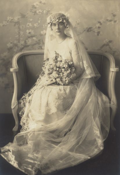 Full-length portrait of Alma Catherine Schmidt, seated, (1894-1989) on the day of her wedding to William F. Petersen M.D. Her waist-length veil is secured by a wreath of flowers, and she holds a large bouquet of sweet pea and lily of the valley flowers. The train of her full-length dress is arranged on the floor in front of her. The painted backdrop features branches with large flowers.