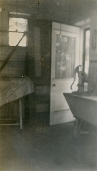 A large sink and hand water pump stand in a corner near a door of the laundry room at Black Point. The laundry was located in the "kitchen building" (demolished 1946) which stood adjacent to the main house.