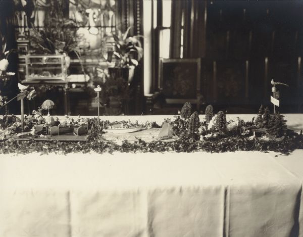 Six scenes of miniature landscapes and interiors comprise a tablescape honoring six decades in the life of Emma Seipp Schmidt on the occasion of her sixtieth birthday. A poem glued to the reverse of the photograph explains the scenes. In this photograph are, from left to right, scenes IV, V, and VI. At left (the 1900's), the Schmidt family enjoys an evening at their home at 3328 South Michigan Avenue, Chicago. In the center (the 1910's), the steamer <i>Loreley</i> is depicted carrying family and guests across Geneva Lake to Black Point Estate. The family's sailboat, <i>Black Point</i>, is also seen. At far right (the 1920's), the Schmidt family enjoys a summer day at the estate. Real foliage and flowers are mingled with artificial trees of loofah and other materials to form the landscape, which is completed with miniature furniture, lamps, dolls and toy animals. In the background is a large stained glass window, live plants and an aquarium on a stand.