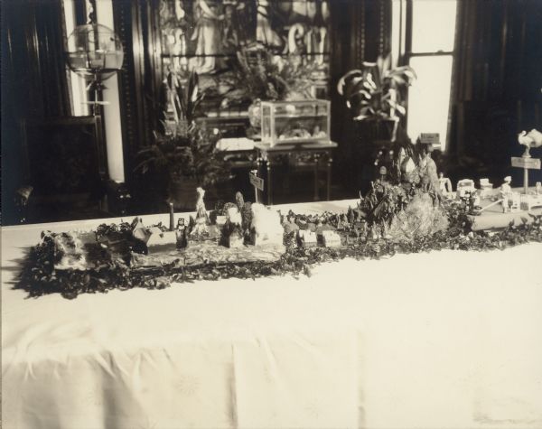 Six scenes of miniature landscapes and interiors comprise a tablescape honoring six decades in the life of Emma Seipp Schmidt on the occasion of her sixtieth birthday. A poem glued to the reverse of the photographs explain the scenes. In this photograph are, from left to right, scenes I, II, and III. At left, scene I (the 1870's) depicts the Great Chicago Fire of 1871. During the 1880's, Emma, with her mother and sisters, spent several years living in Wiesbaden, Germany. The center scene recalls their time in Germany, with a village of small wooden houses, carved wooden figures and trees all from the Erzgebirge region of Germany. At far right (the 1890's), a stork on a pole watches over the nursery in the Schmidt home at e3328 South Michigan Avenue, Chicago. The nursery has miniature beds, a dresser, chairs and a sewing machine. There are dolls representing the three Schmidt children and their nanny. Real foliage and flowers complete the scenes. In the background is large stained glass window, an aquarium on a stand, live plants and a birdcage.