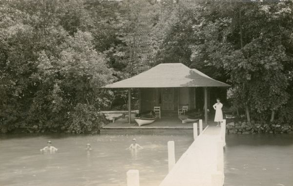 View from end of pier towards three boys posing in the water of Geneva Lake in front of the boathouse at Black Point Estate. A woman observes from the narrow pier near the shoreline. Two boats rest on the broad, deep porch. Written next to the photograph is "In the 1930's. Elly who could not swim watching Edward — Conrad and Nathan."
