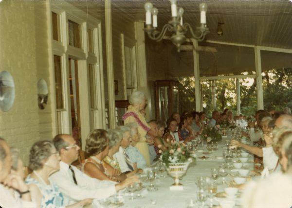 Alma Schmidt Petersen, wearing a summer dress and flower lei, stands at a long table on the dining porch of the main house at Black Point Estate surrounded by seated family members and friends. She was being honored on the occasion of her eightieth birthday.