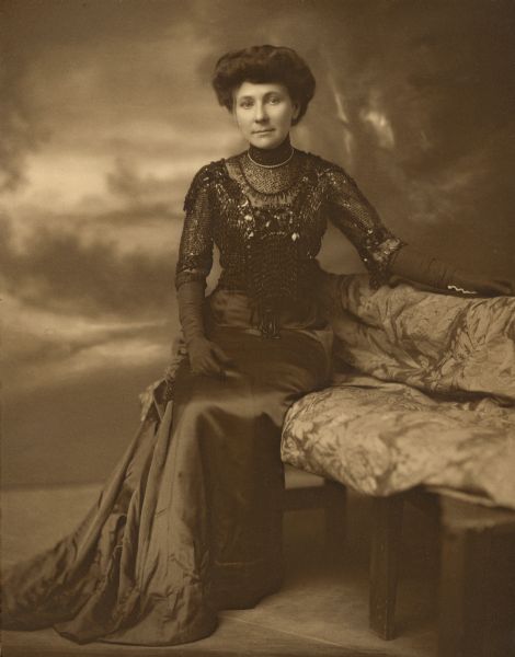 Seated, full-length portrait in front of a painted backdrop of Alma Seipp (1876-1966), daughter of prominent Chicago brewer Conrad Seipp. She is seated on a love seat and wears a full-length gown with a high sheen skirt and extensively beaded bodice. She has elbow length dark gloves and a single strand of pearls.