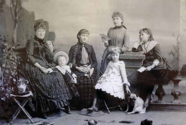 Group studio portrait in front of a painted background. Catherine Orb Seipp, seated at left, poses with her children. From left: Conrad, Clara, Emma (standing), Alma and Elsa. Alma is holding the collar of a pug puppy. In the custom of the time, Conrad is wearing a velvet dress with large lace collar and cuffs.