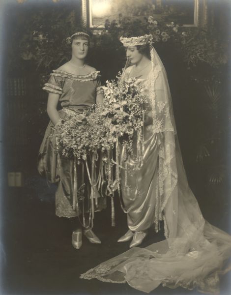 Bride Catherine Bartholomay, right, poses with her sister Elsa.  Catherine wears an ankle length gown with long veil. Both women hold large bouquets of fresh flowers festooned with ribbons.