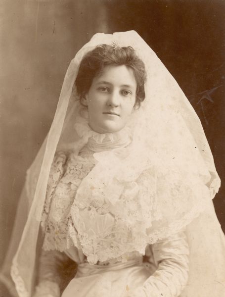 Waist-up standing portrait of Elsa Seipp Madlener on her wedding day. She is wearing a long-sleeved dress with a high collar, lace bodice and a lace veil which is held in place with a jeweled hairpin.