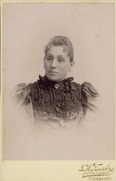 Quarter-length studio portrait of Maria Best Pabst (1842-1906). She is wearing a dress with leg of mutton sleeves and ornate embroidery. The daughter of successful Milwaukee brewer Phillip Best, Maria married Captain Frederick Pabst in 1862. Together they had ten children, only five of whom survived to adulthood. Pabst went into partnership with his father-in-law in 1863 and eventually owned what would become the Pabst Brewery.