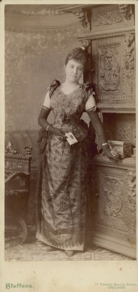 Full-length portrait of Emma Huck Seipp (1855-1891). She is standing and wearing a floor length sleeveless gown and opera length gloves. Emma was the first wife of William Conrad Seipp, whose father, Chicago brewer Conrad Seipp, built Black Point Estate.
