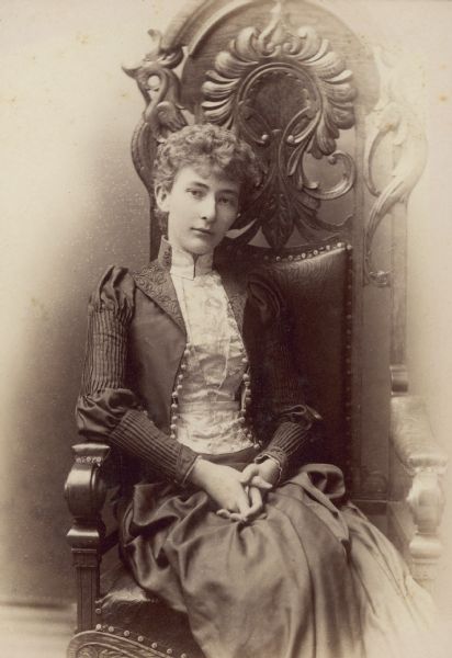 Three-quarter length studio portrait of Emma Seipp, daughter of Chicago brewer Conrad Seipp. She is wearing a short jacket over a blouse and skirt. She is seated on a chair with an ornately carved wooden back.