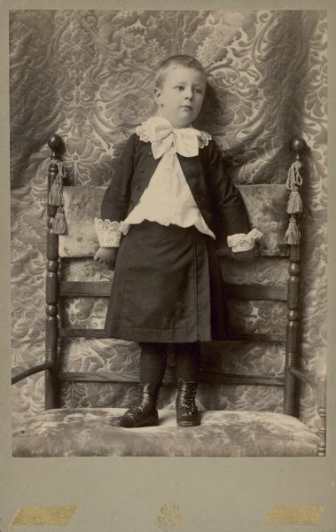 A young Conrad Seipp Jr. stands on an oversized chair for a full-length studio portrait. He is wearing a skirt over long stockings, a jacket and white blouse with a large bow and lace collar and cuffs. He was the youngest child of Chicago brewer Conrad Seipp.