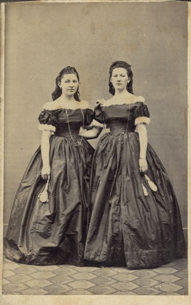 Sisters Margarethe, left, and Catherine Orb posing for a full-length, standing portrait wearing matching dark floor-length gowns with fur-trimmed short sleeves and necklines. Both are wearing earrings and a long watch chain and watch. 