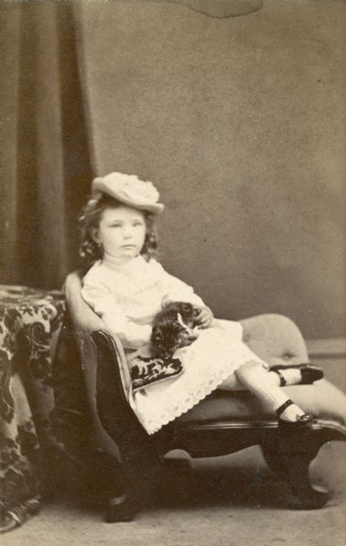 Emma Seipp poses for her portrait seated on a chair holding a kitten and a small pillow in her lap. Emma is wearing a short dress with lace trim, a hat, and long stockings with shoes with ankle straps.