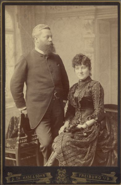 August Streb leans against a chair as he poses next to his wife, Margarthe Orb Streb, who is seated. He is wearing a suit and a watch chain. Margarethe is wearing an elaborately detailed gown and holds a fan. The portrait was taken in Freiburg, Germany and may be a wedding portrait.