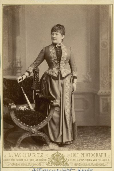 Full-length studio portrait in front of a painted backdrop of Catherine Orb Seipp (1846-1920), wife of Chicago brewer Conrad Seipp. She is standing and wearing a floor-length tailored dress with elaborate embroidered trim, a comb in her hair, and a large anchor-shaped brooch at her neck. Her right hand, with the strap of a large hand fan draped over her wrist, rests on the back of a chair. The chair has a carved frame, and a small bouquet of flowers rests on the seat.