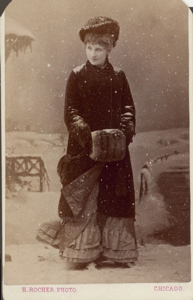 Studio portrait of an unidentified woman dressed in a winter coat, hat and muff. Her coat has been turned back to reveal the ruffles on the bottom of her skirt. There is faux snow on her clothing and on the floor. The photograph has been altered to simulate falling snow. A painted winter backdrop completes the effect.