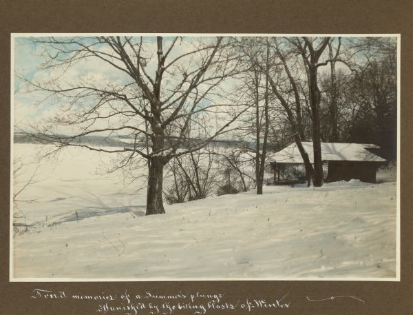 A small wood frame boathouse with a large front porch stands at the snow covered shore of frozen Geneva Lake at Black Point. Evergreens and bare deciduous trees frame the view. The photographer has written below the photograph: "Fond memories of a Summer's plunge Banished by the biting blasts of Winter."