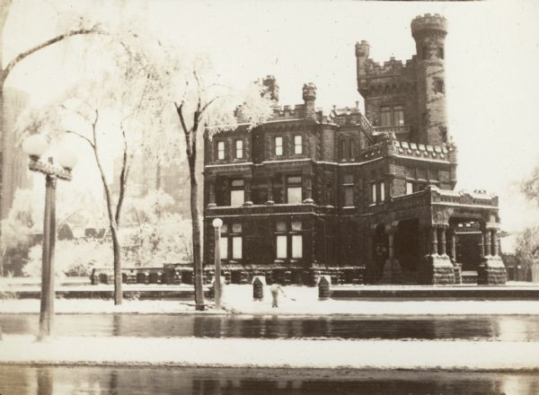 View from across street towards a worker shoveling a sidewalk in front of the Potter Palmer mansion on North Lake Shore Drive. The three-story granite faced house has crenellated towers and balustrades and a large entry porch. The trees are ice covered and there is snow on the ground.