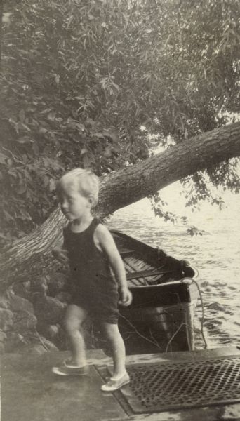 Conrad Petersen steps off the pier onto the shore at Black Point Estate.  A small boat is moored to the shore in the background under a tree trunk leaning over the water of Geneva Lake.