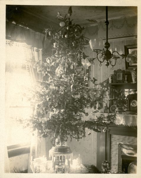 Christmas tree in the home of Edward Petersen decorated with a wide assortment of ornaments and tinsel garland. It is standing on a low table in front of a parlor window. The tree is in a mechanical, rotating stand which itself is incorporated in a winter scene under the tree. There is a transitional ceiling chandelier with both gas and electric arms. The fireplace mantle is crowded with vases, a clock, and a picture frame.