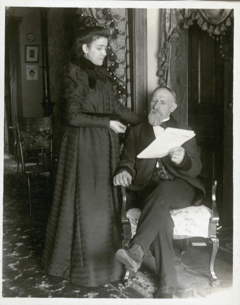 Edward Petersen poses, seated in a chair, and his wife stands next to him on the left. They appear to be reading papers which Edward is holding. Behind them is an open doorway to the left and a closed door on the right. There are tasseled draperies in the doorways.