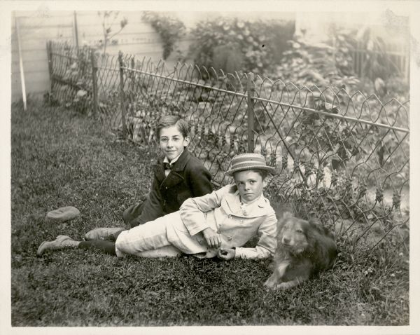 William F. Petersen, rear, poses on the lawn with an unidentified boy and a dog. Behind them is a low, fancy metal wire fence which encloses a garden near the side of a building. The boys are well-dressed; William's cap is on the ground and his friend is wearing a straw boater.